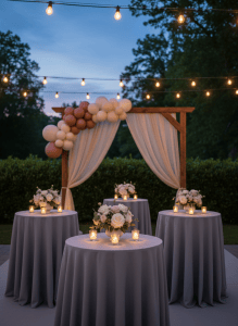 An intimate outdoor garden event setup in Northern Virginia at twilight, with a small cluster of round cocktail tables draped in smooth, dove-gray linens. Each table features a low arrangement of white and blush flowers in a simple ceramic vase, surrounded by glass votives. In the background, a custom backdrop structure with flowing neutral fabric panels and a restrained balloon installation in soft, matte tones frames the space. Subtle string lights overhead and the last traces of blue evening sky provide layered, ambient lighting, creating gentle reflections on the glassware. Photographic realism, shot from a slightly elevated, wide-angle perspective, balances the tables in the foreground with the backdrop in the distance, conveying a serene, sophisticated, and curated atmosphere for upscale event planning.